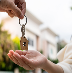 Close-up of two hands exchanging a keychain shaped like a small wooden house, symbolizing a real estate transaction or new home ownership. Blurred background shows suburban homes and greenery.